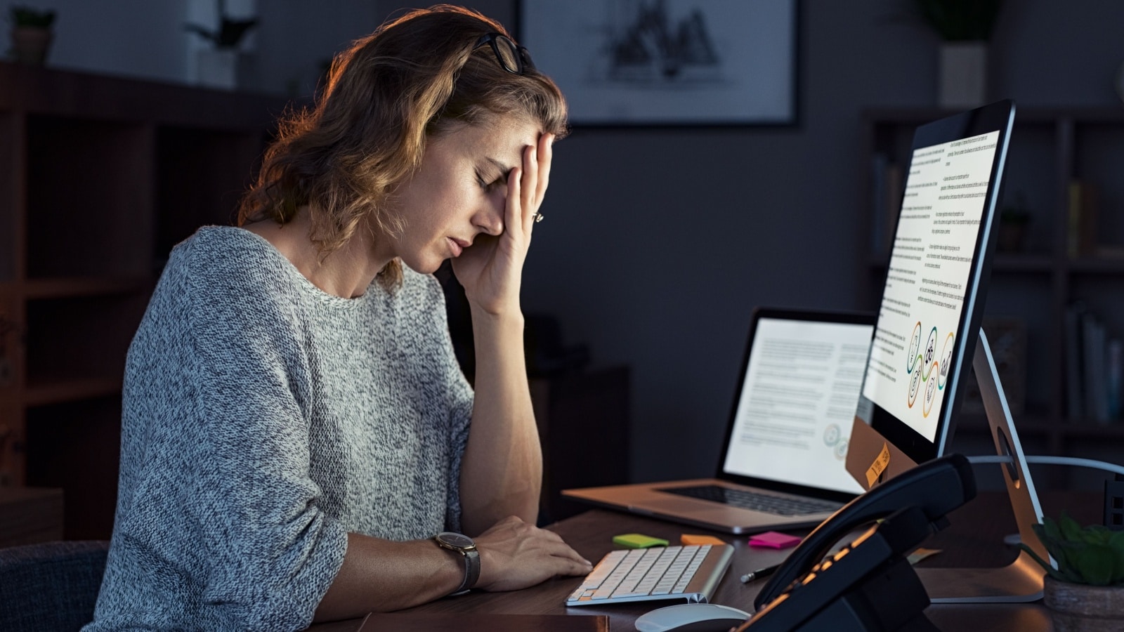 A tired looking woman sits in front of a computer with her hand on her head.