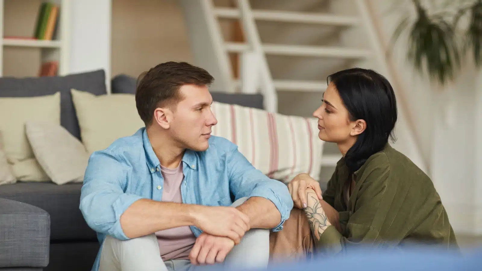 A couple sits on the floor in front of the couch looking at each other with serious expressions as though they're deep in a serious conversation.