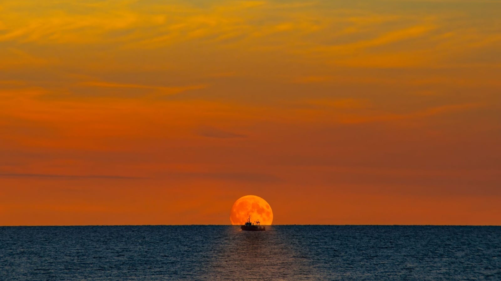 A fishing boat on the horizon at sunset looks tiny compared to the vast ocean. 