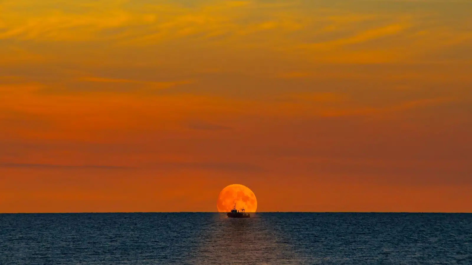 A fishing boat on the horizon at sunset looks tiny compared to the vast ocean.