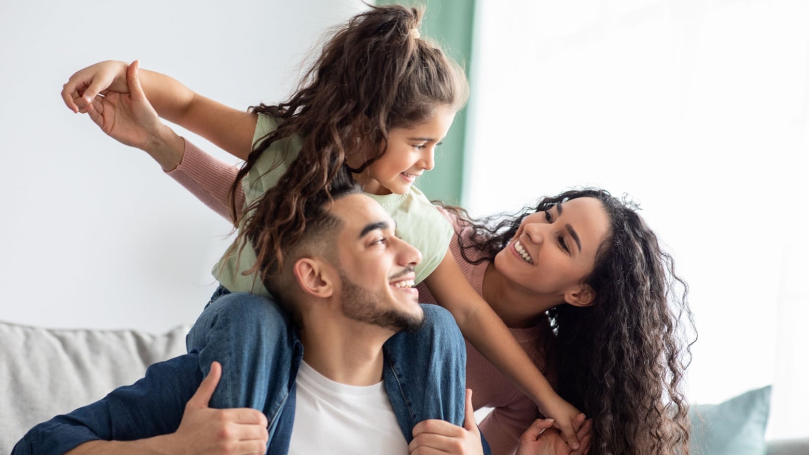 A happy family sits on the couch. The daughter is on the father's shoulders.