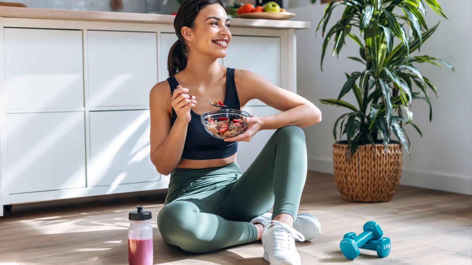 A woman sits on the floor in work out clothes eating a healthy meal to represent healthy habits.