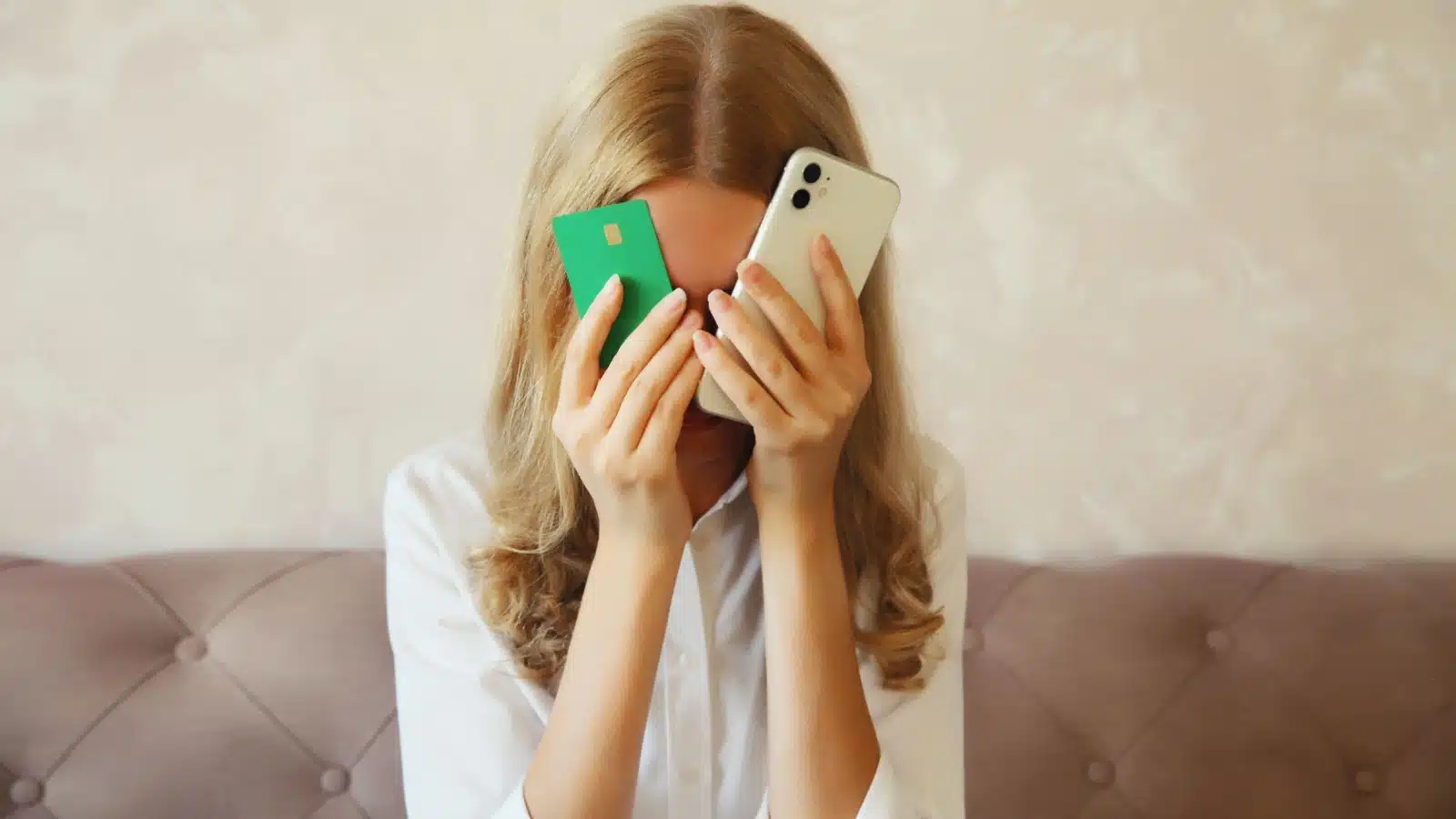 An upset woman hides her face behind a credit card and mobile phone. She's overspending, but you can learn how to stop spending money.