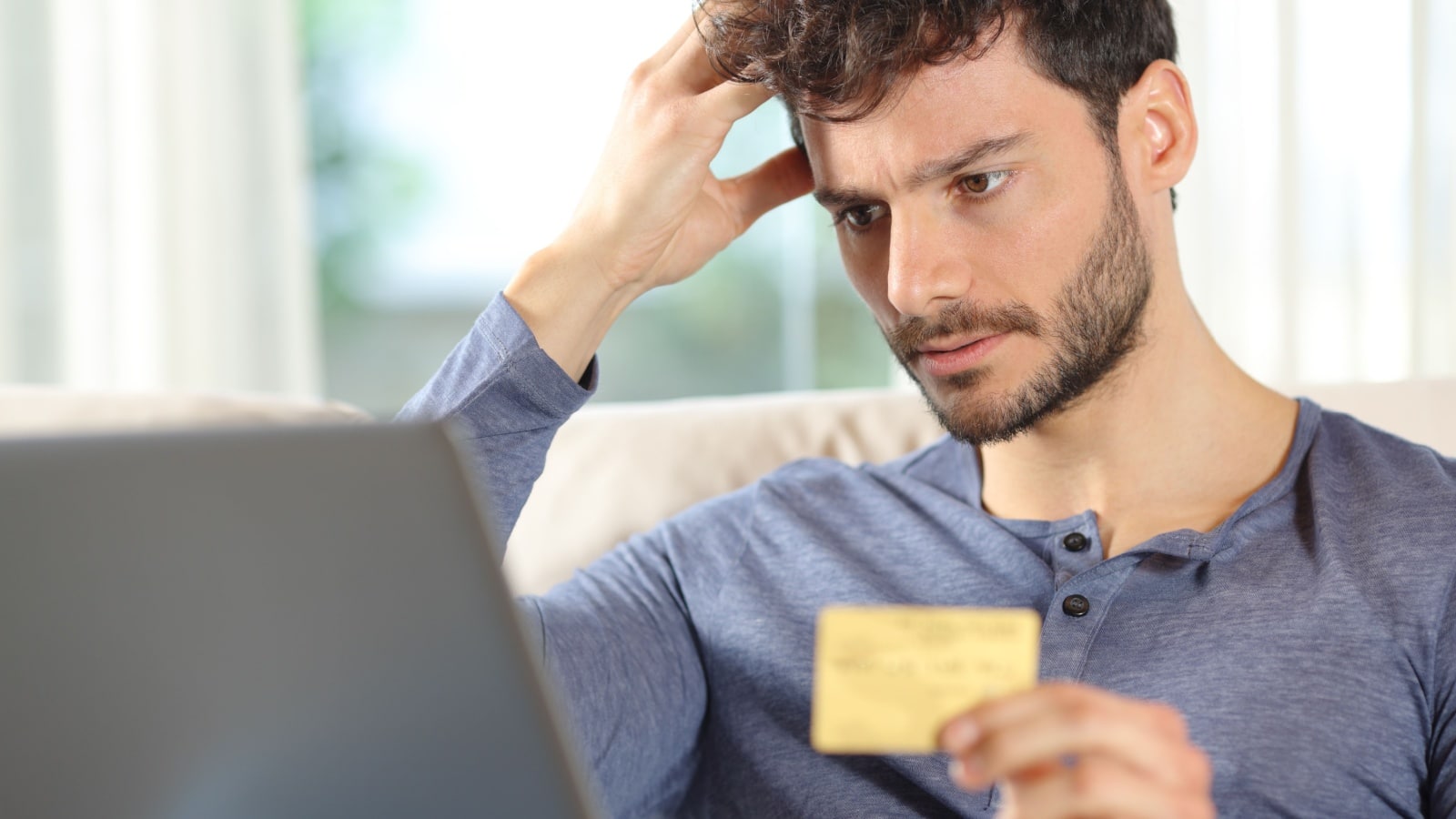 An anxious man looks at his laptop with a credit card in hand, to highlight that he's spent too much money.
