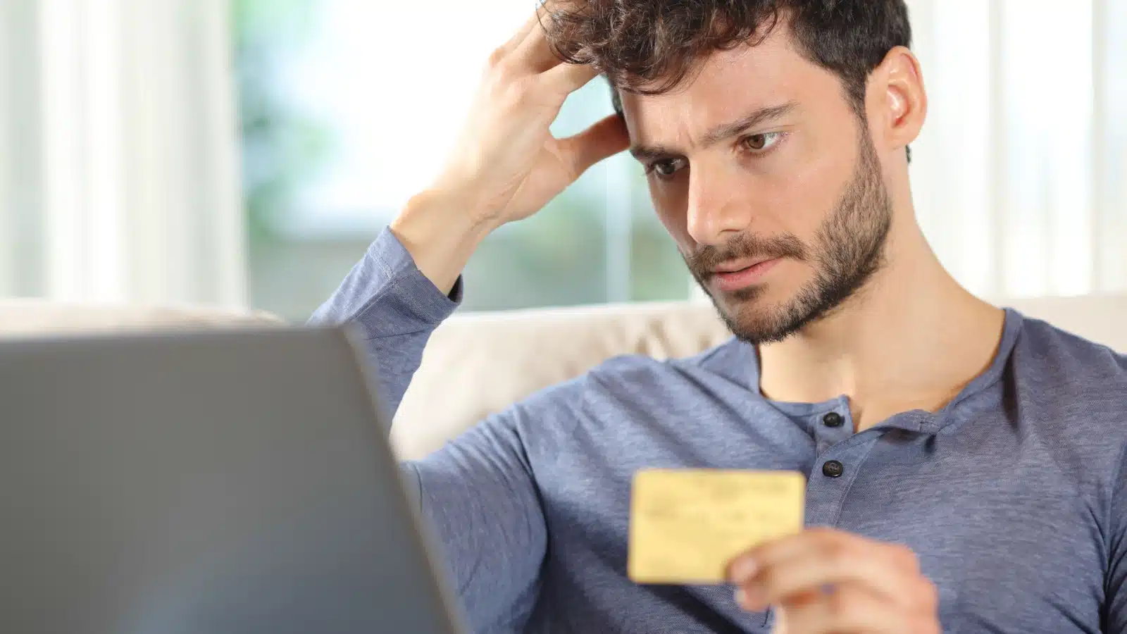 An anxious man looks at his laptop with a credit card in hand, to highlight that he's spent too much money.