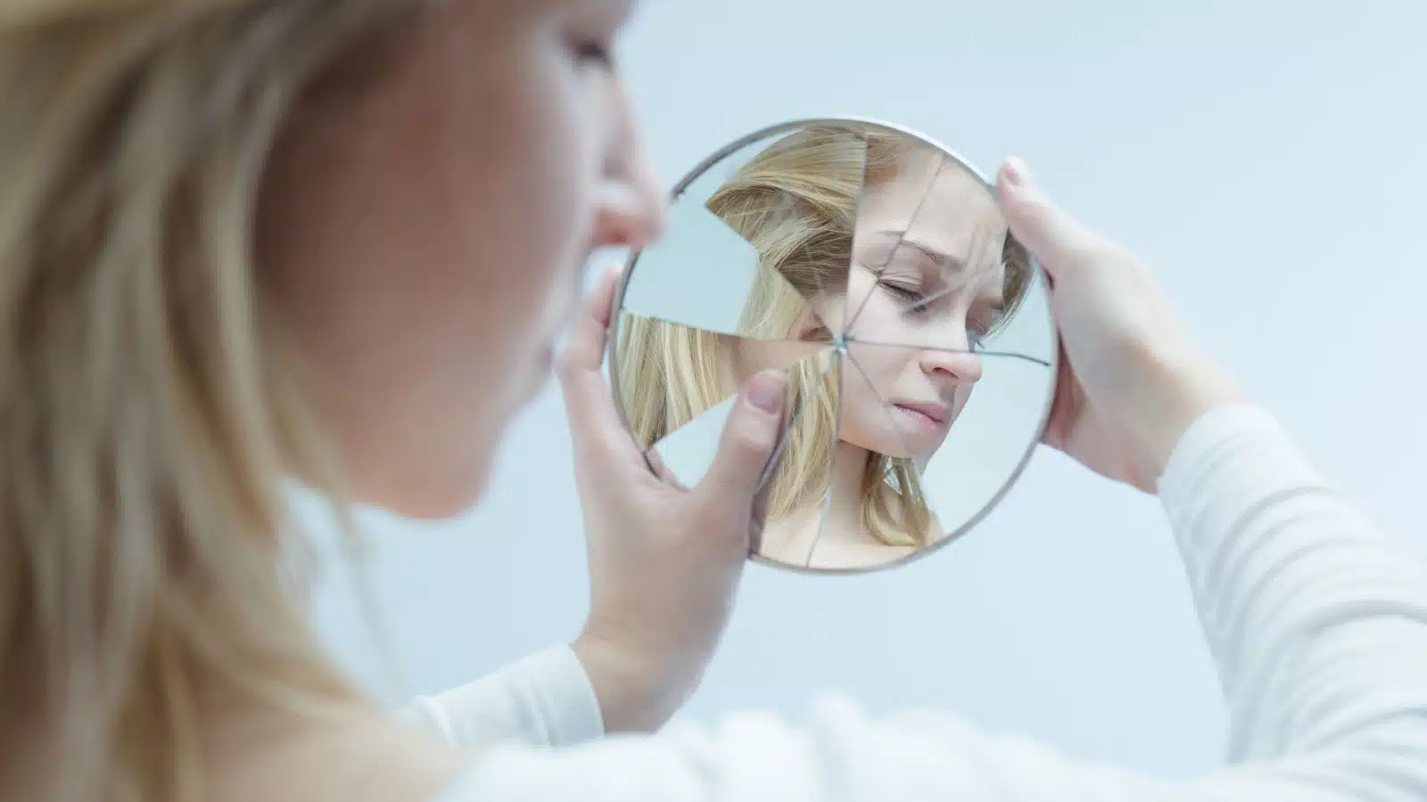 A sad woman looks away from a shattered mirror image of herself, to represent low self-esteem.