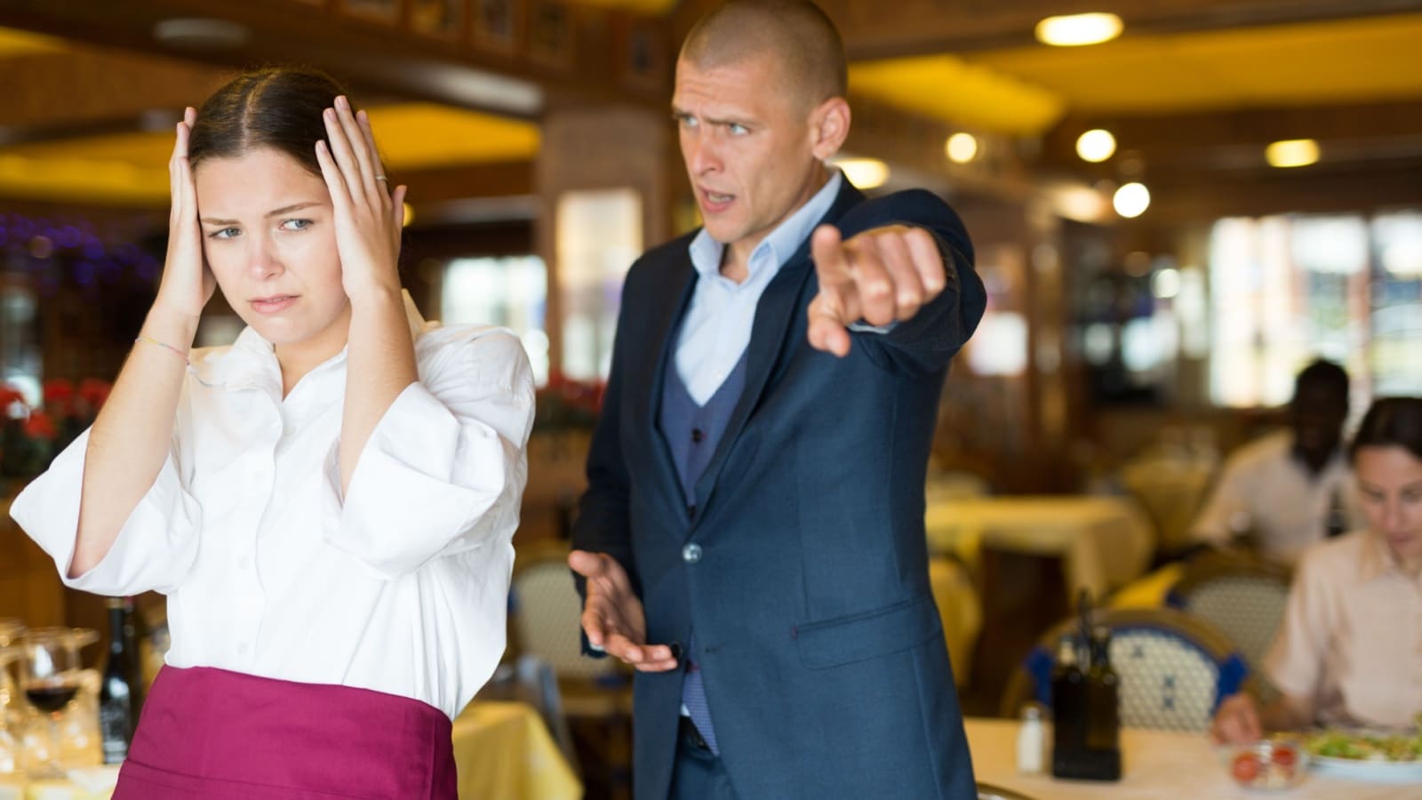 An angry man yells at a server in a restaurant while she covers her ears.