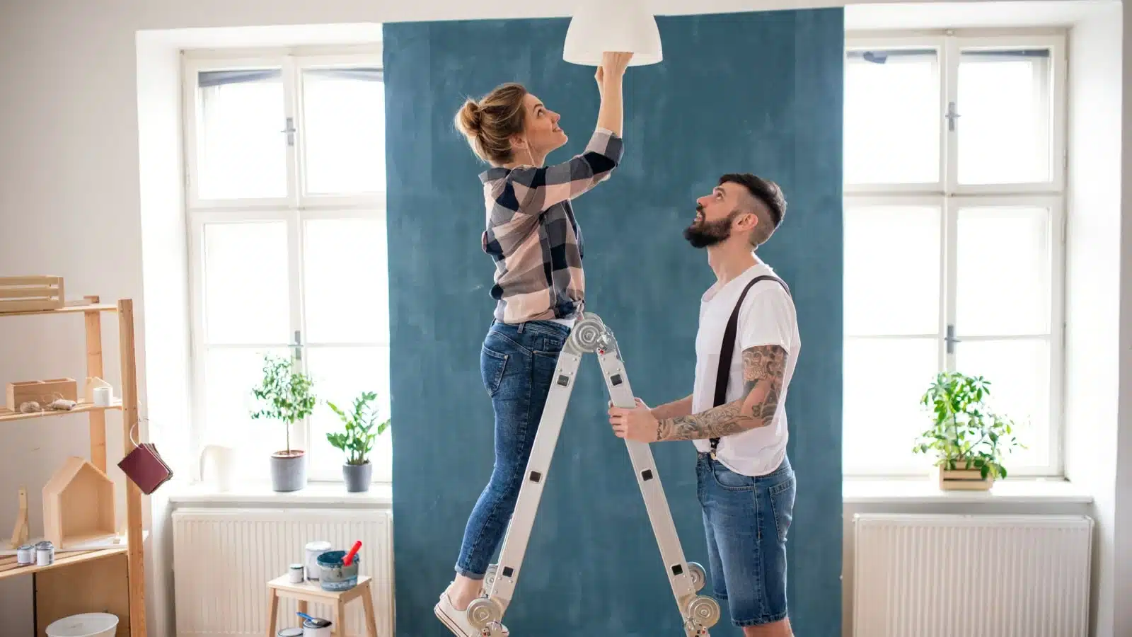 A couple works as a team to change a lightbulb. He holds the ladder while she changes the bulb.