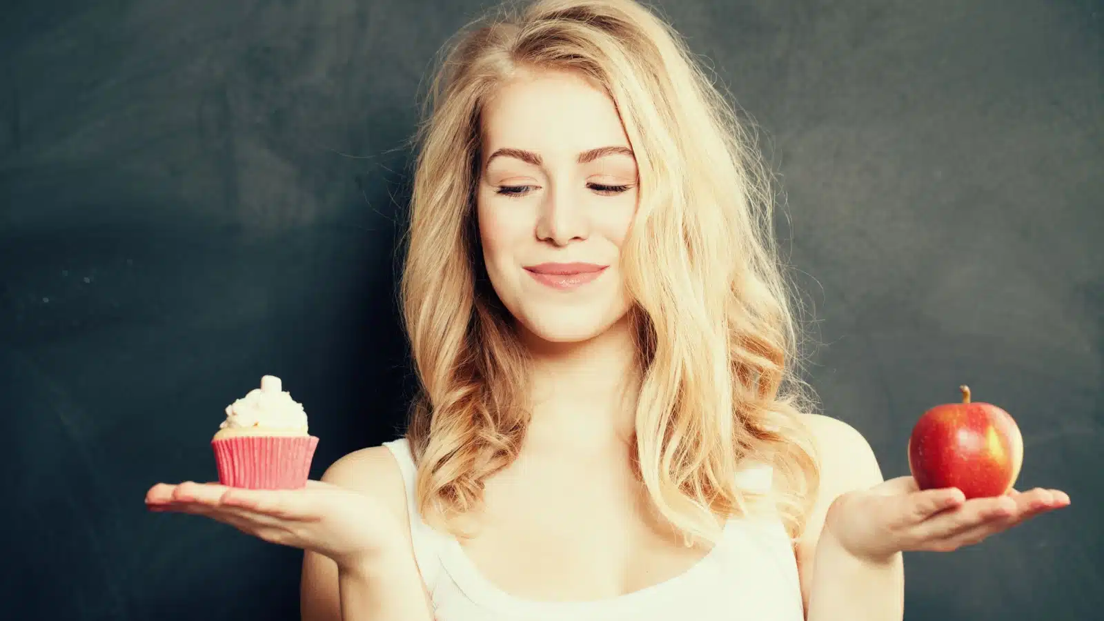 A woman holds a cupcake in one hand and an apple in the other. She's looking at the cupcake as if it's tempting her.