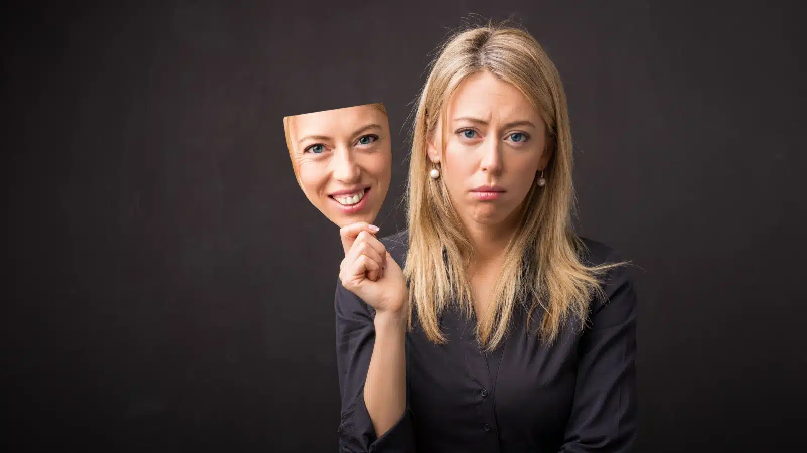 A sad woman holds a mask of her face wearing a fake smile to represent toxic positivity.