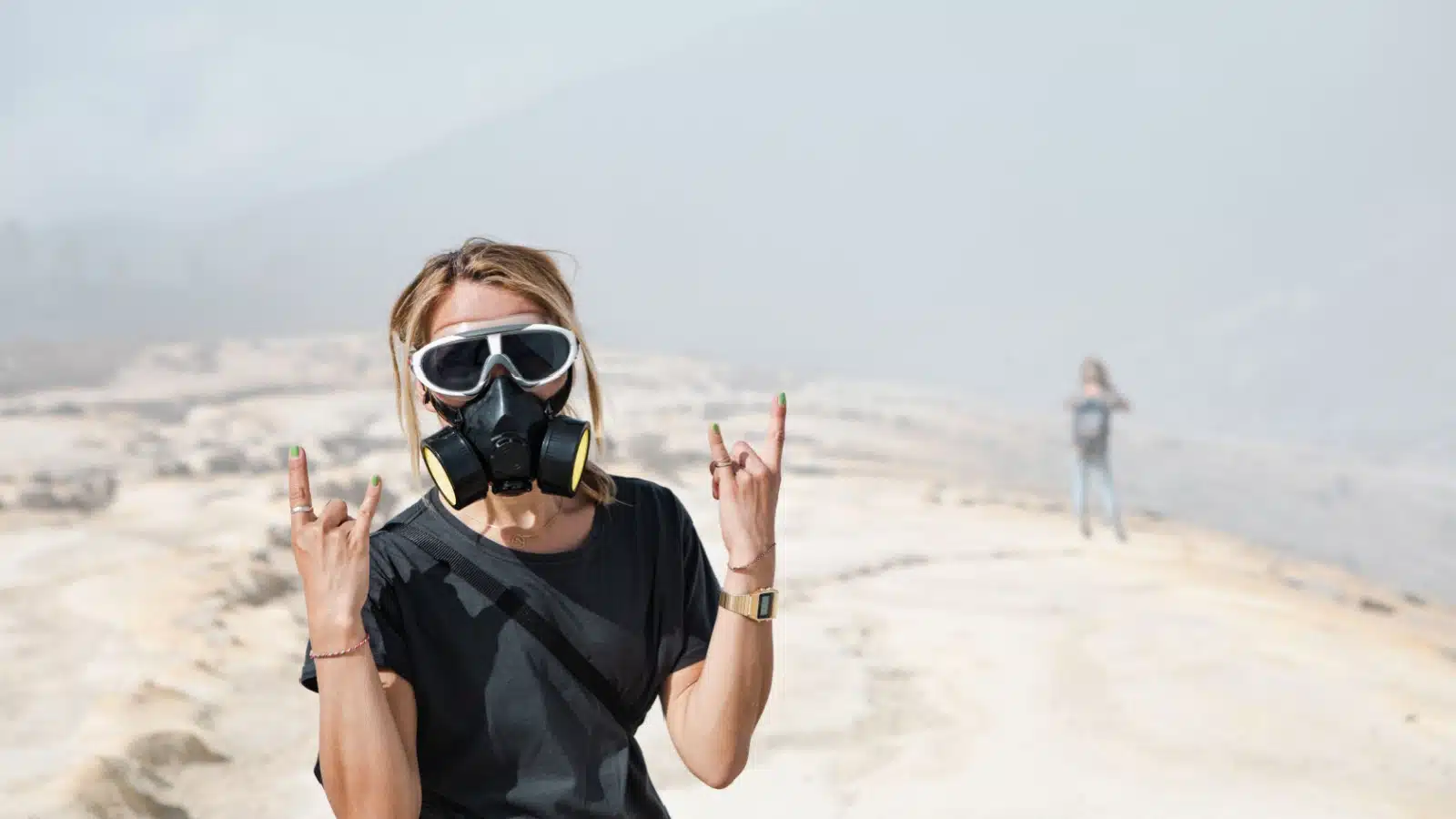 A woman poses with hand gestures indicating she's enjoying herself while wearing a gas mask in a wasteland.