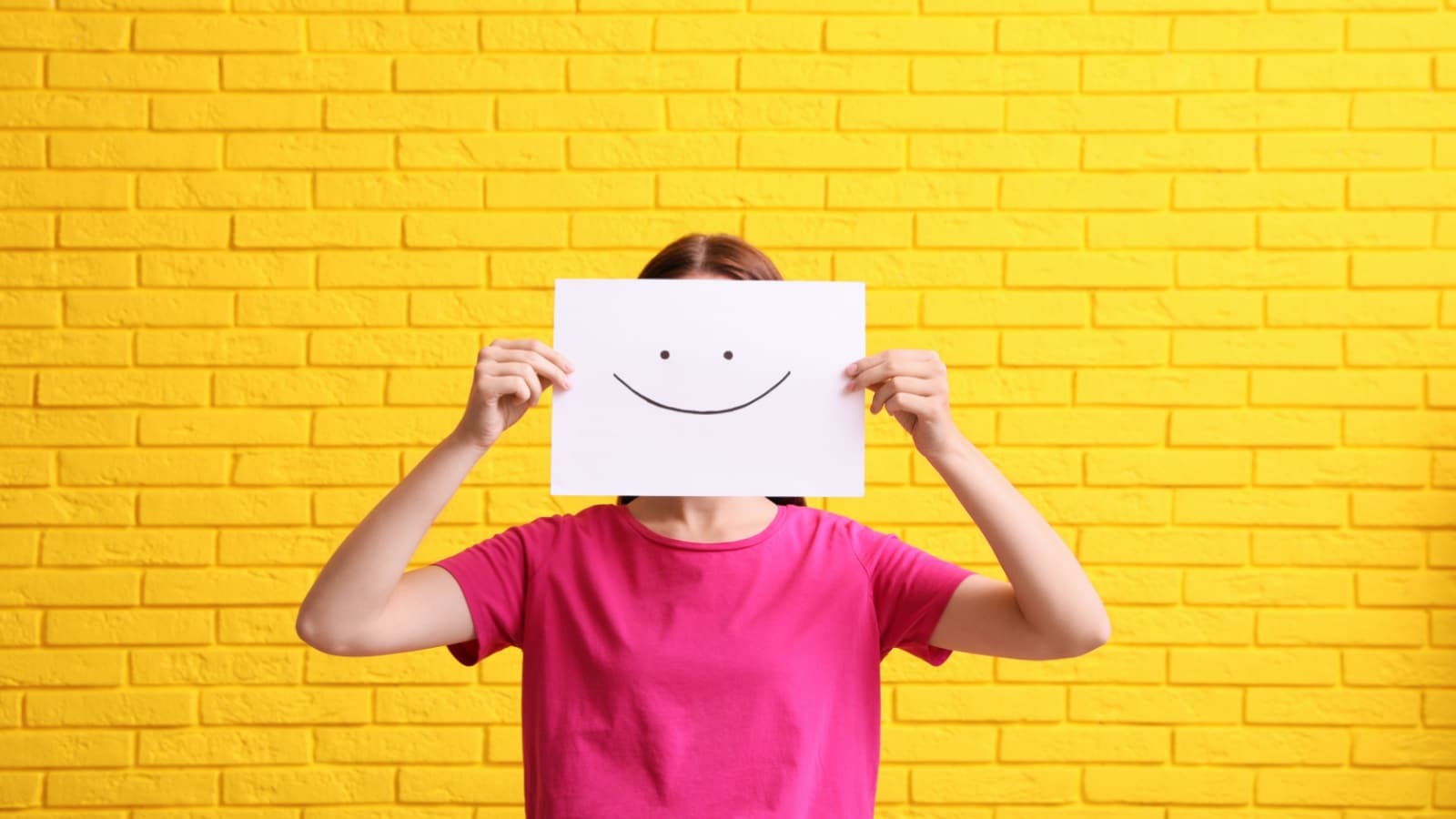 A woman stands in front of a bright yellow brick wall holding a paper sketch of a smile in front of her face to represent toxic positivity.