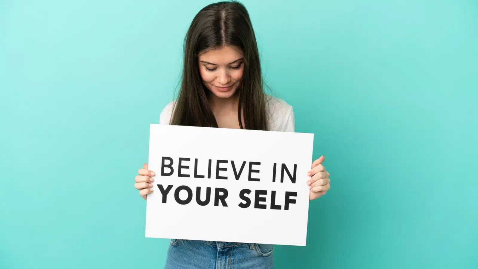 A woman looks down at the sign she's holding which says "believe in yourself" but how do you accomplish that?