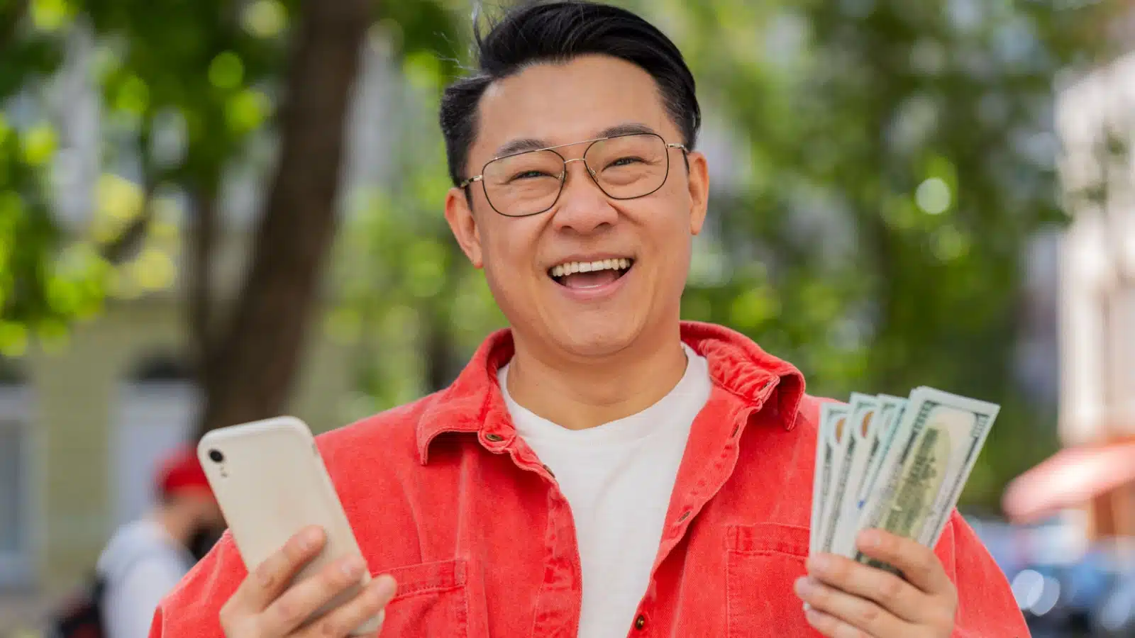 A smiling man holds his cell phone in one hand and a pile of money in the other.