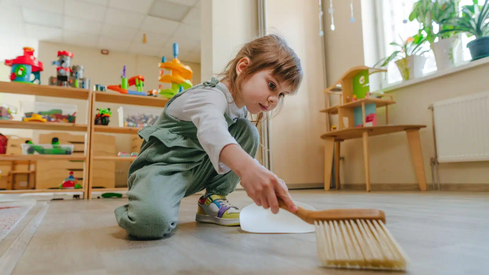 A little girl using a small broom and dustpan to clean, representing how we socialize young girls to clean from an early age.