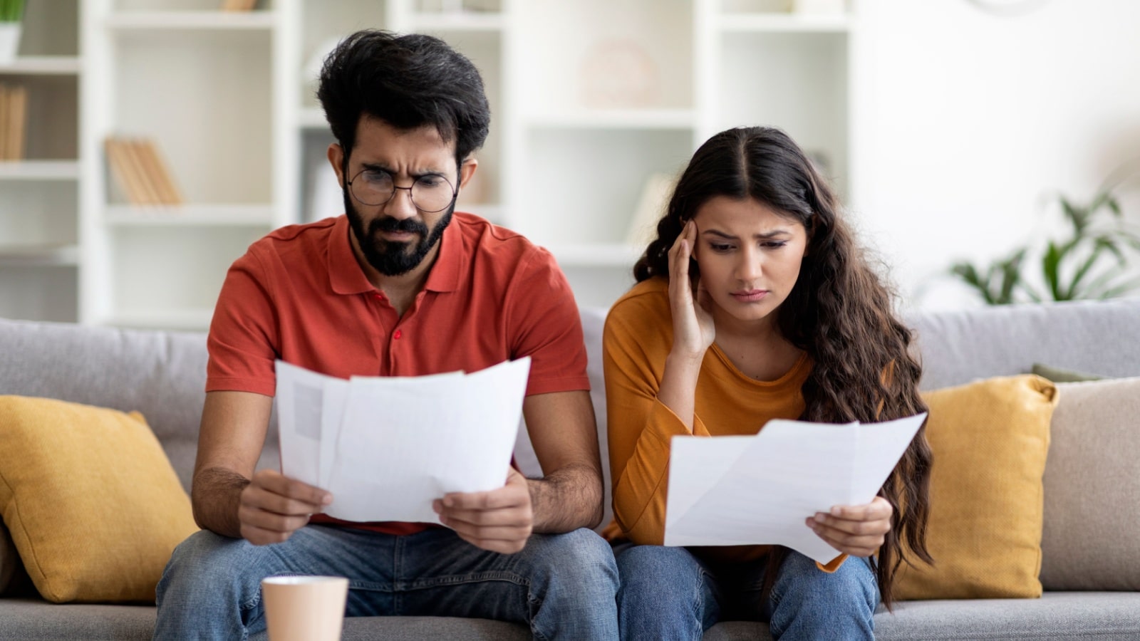 A stressed couple sits on the couch reviewing financial paperwork because they have money problems.
