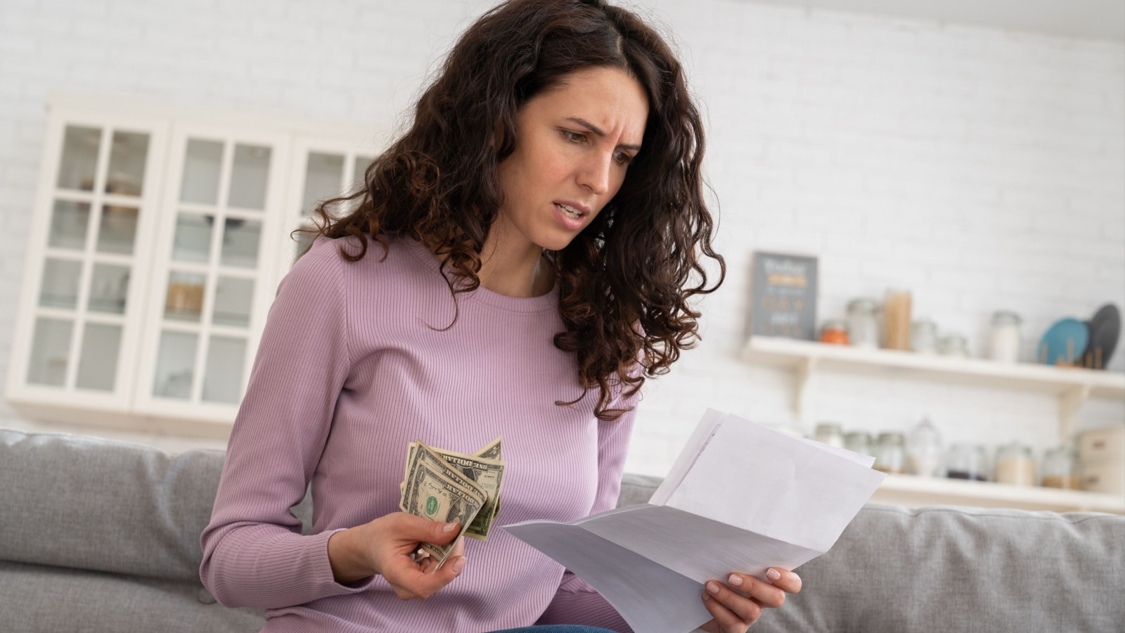 A woman nervously looks at her bills while holding cash, to represent someone who is house poor and can barely keep up with bills.