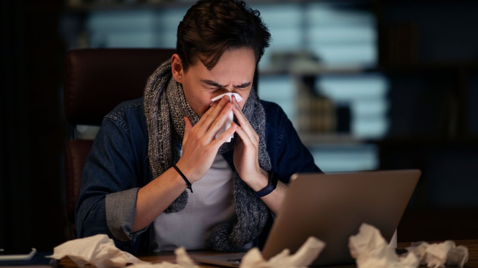 A sick man blows his nose while working at his laptop, to represent someone forced to work when sick.