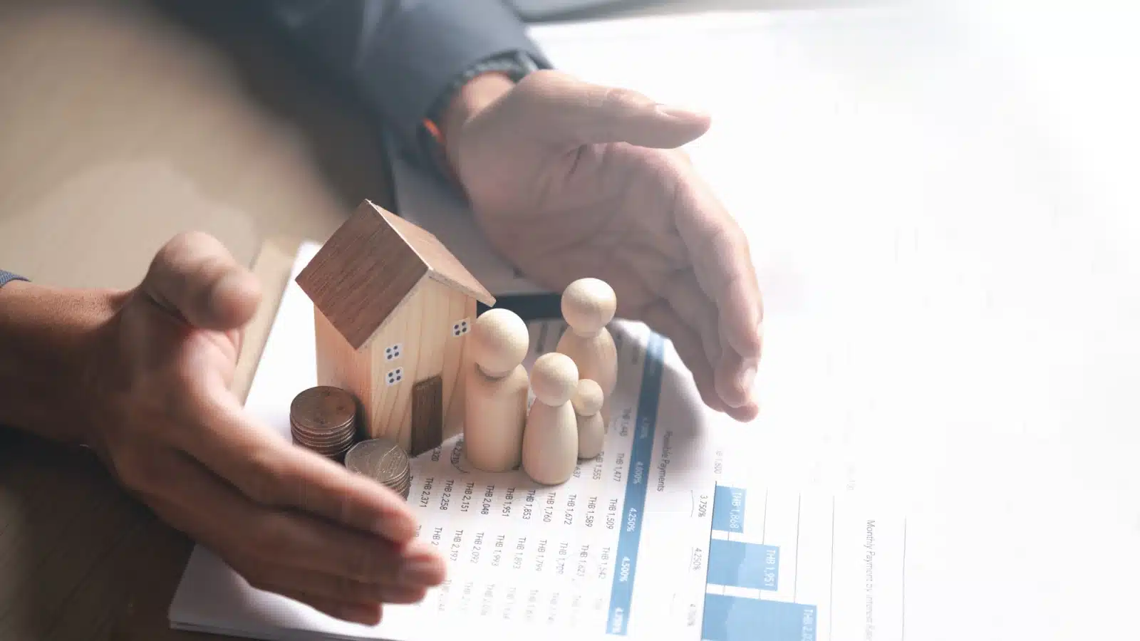 Hands protecting a small model home and the people inside on top of paperwork, to represent a home warranty and home insurance.
