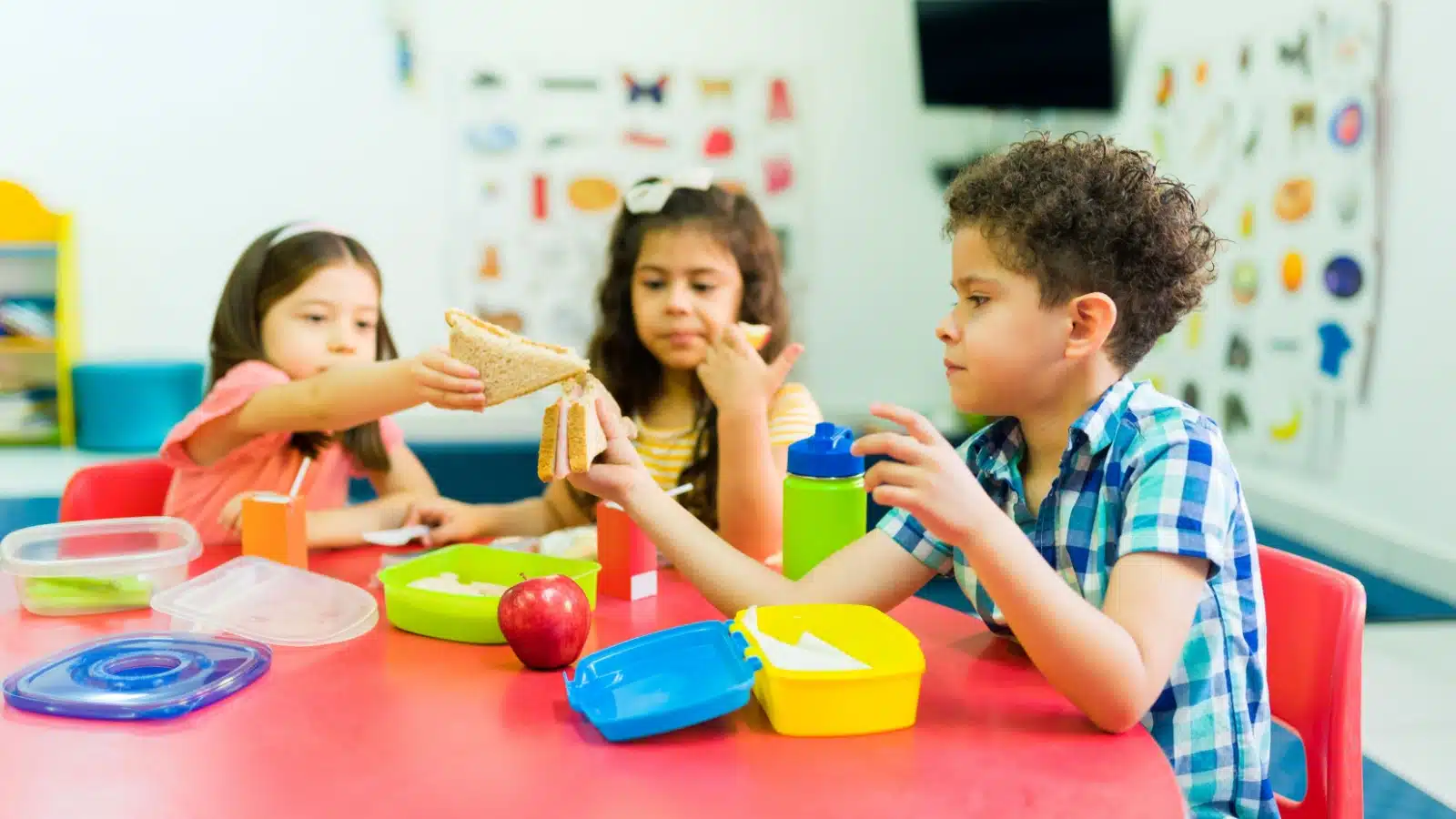 Three young children sharing their lunches at a school table.