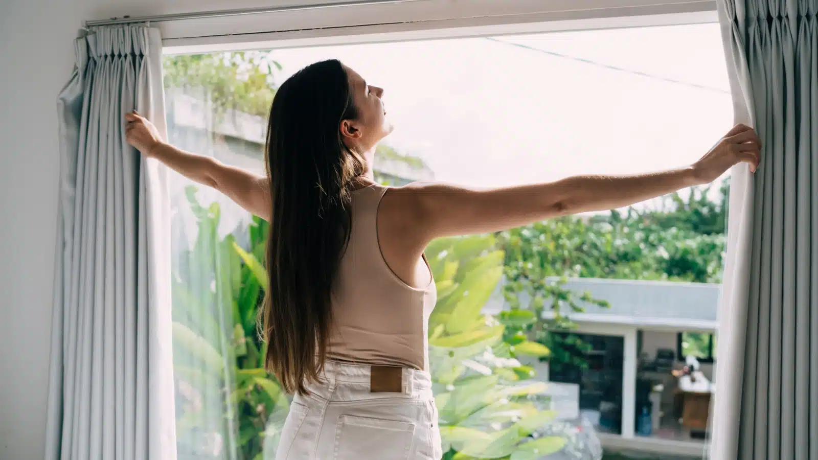 A woman thrusts open her curtains and lets the sun shine on her face, to represent setting an intention for a happy day.