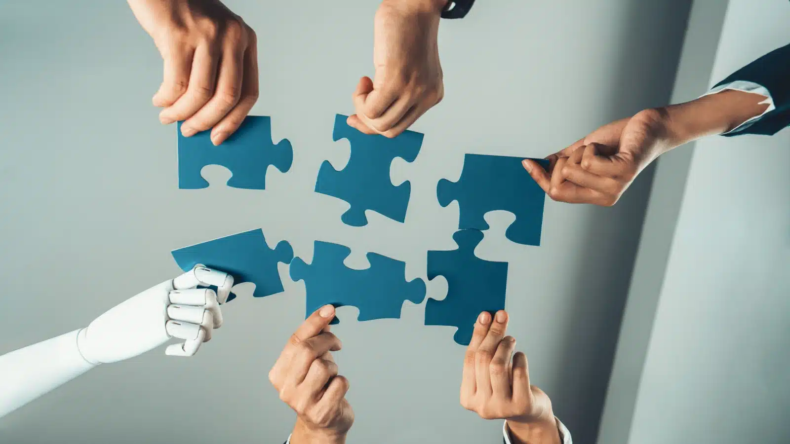 Six hands putting puzzle pieces together. One of them is a robot to highlight the threat of automation against jobs.