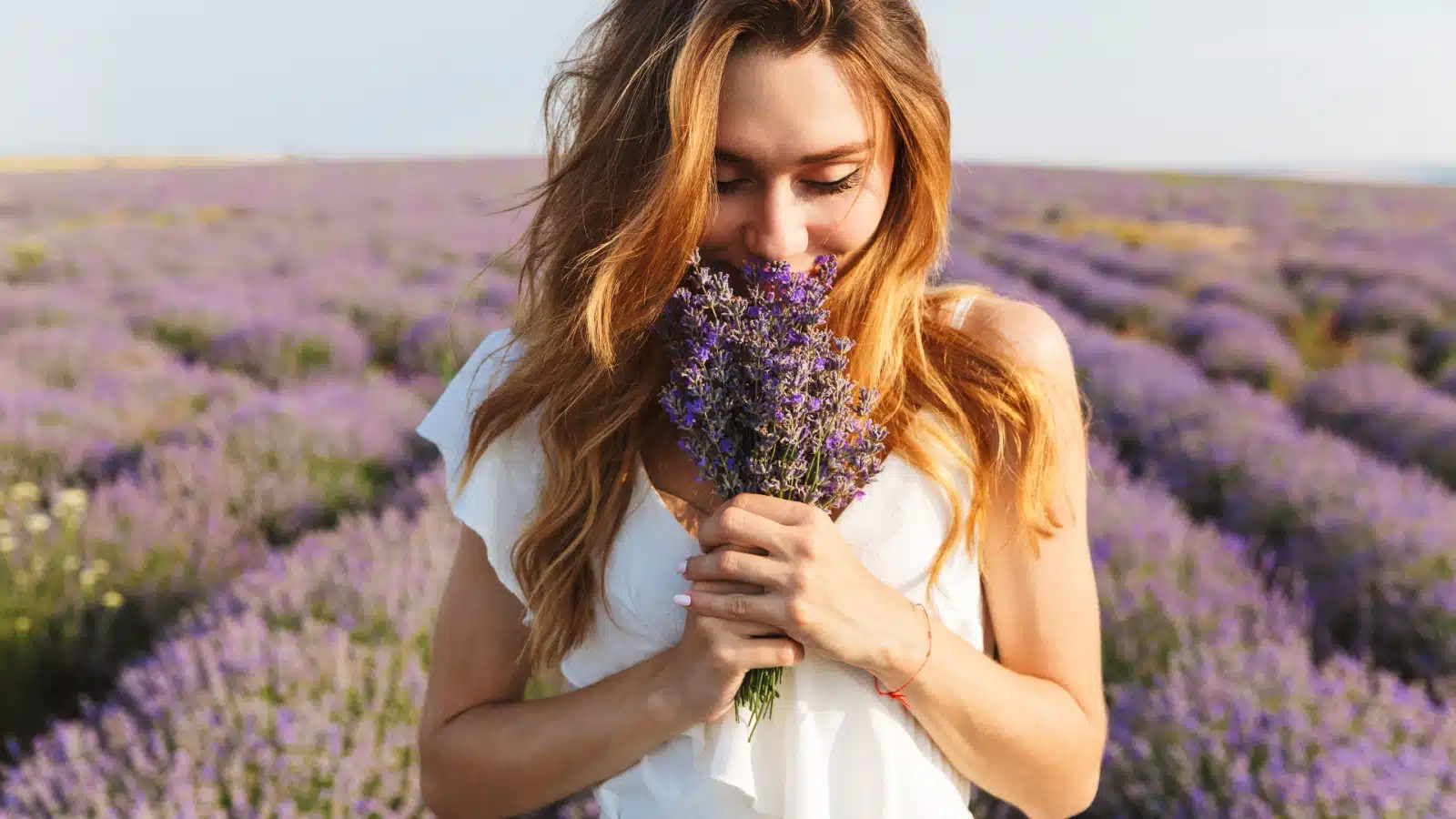 A woman in a field of lavender stops and smells a bunch in her hand.