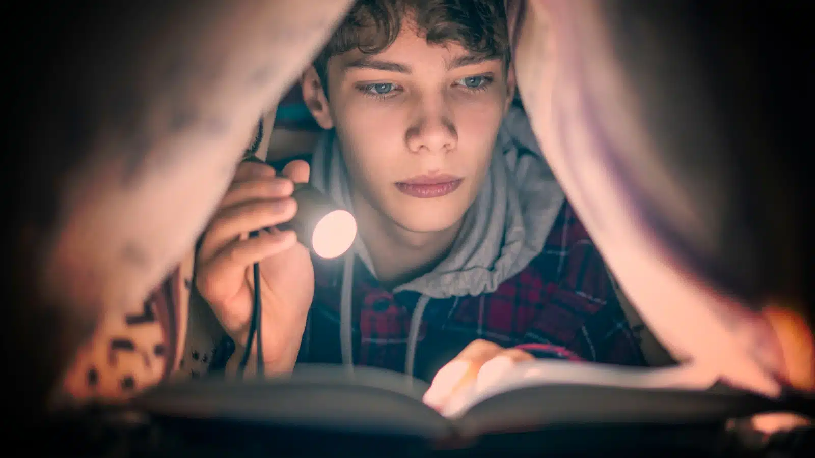 A teenager reading a scary story under a tent by flashlight.
