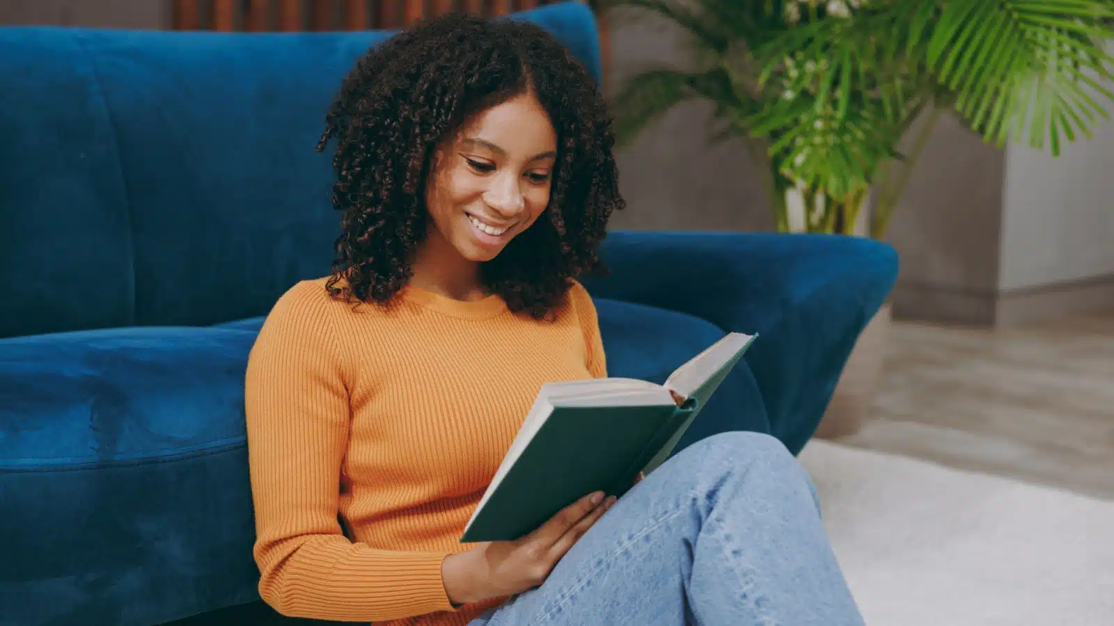 A young woman smiles as she sits on the floor reading a book.