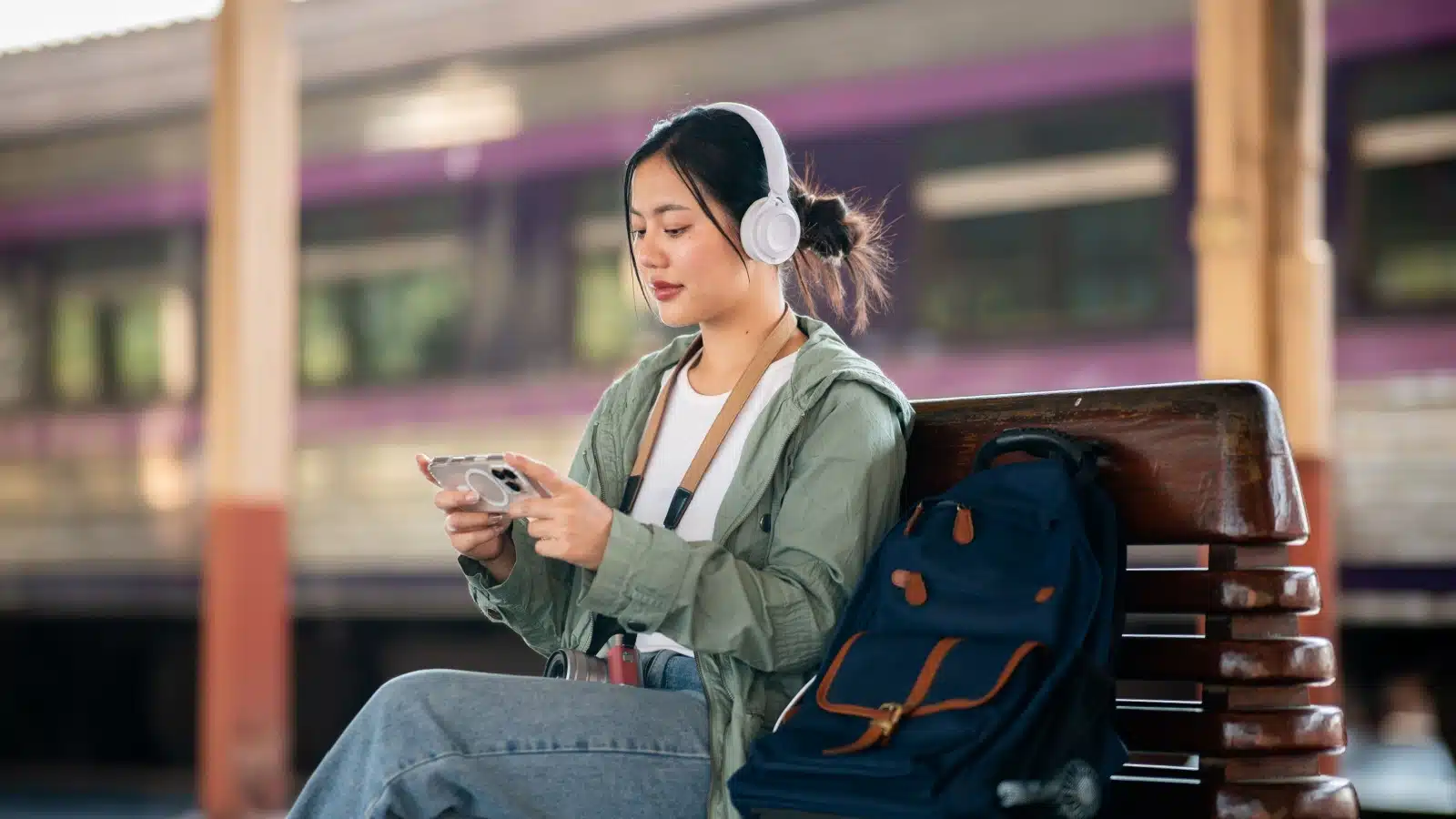 A woman wearing headphones plays on her phone while waiting at the train station.