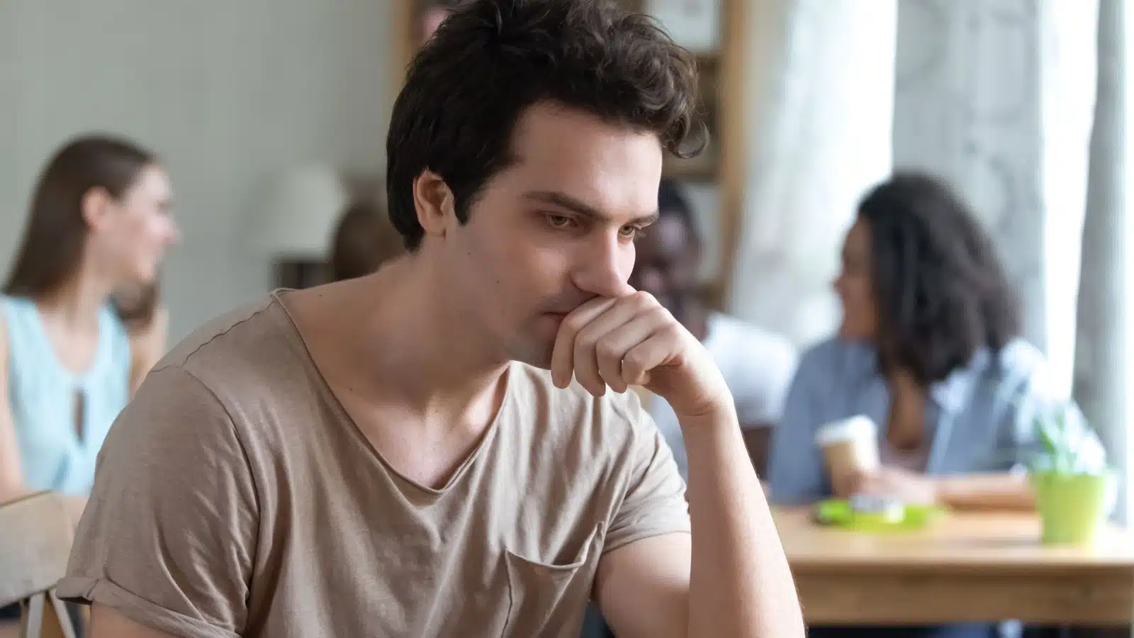 An upset man looks thoughtful as he sits away from a group of friends. He's pondering the behaviors that make people dislike you so he can be a better friend.