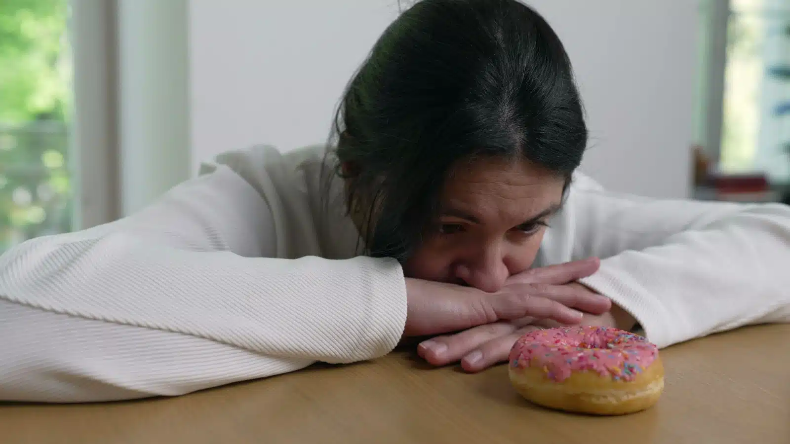 A woman stares longingly at a donut, but doesn't touch it, to represent willpower and delayed gratification.