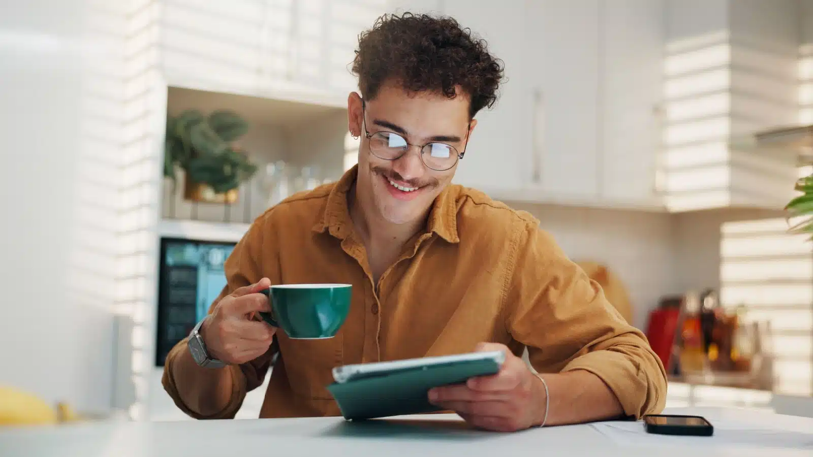 A man smiles as he reads happy news on his tablet while drinking his morning coffee.
