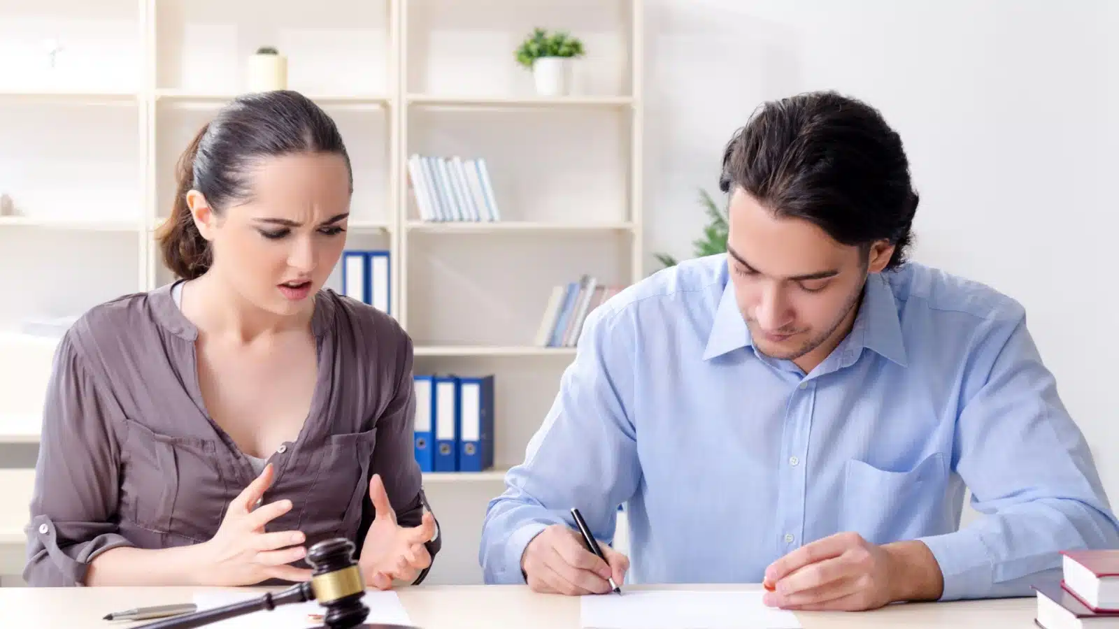 A woman looks upset while her partner happily signs a prenup, to represent the question why do women hate prenups.
