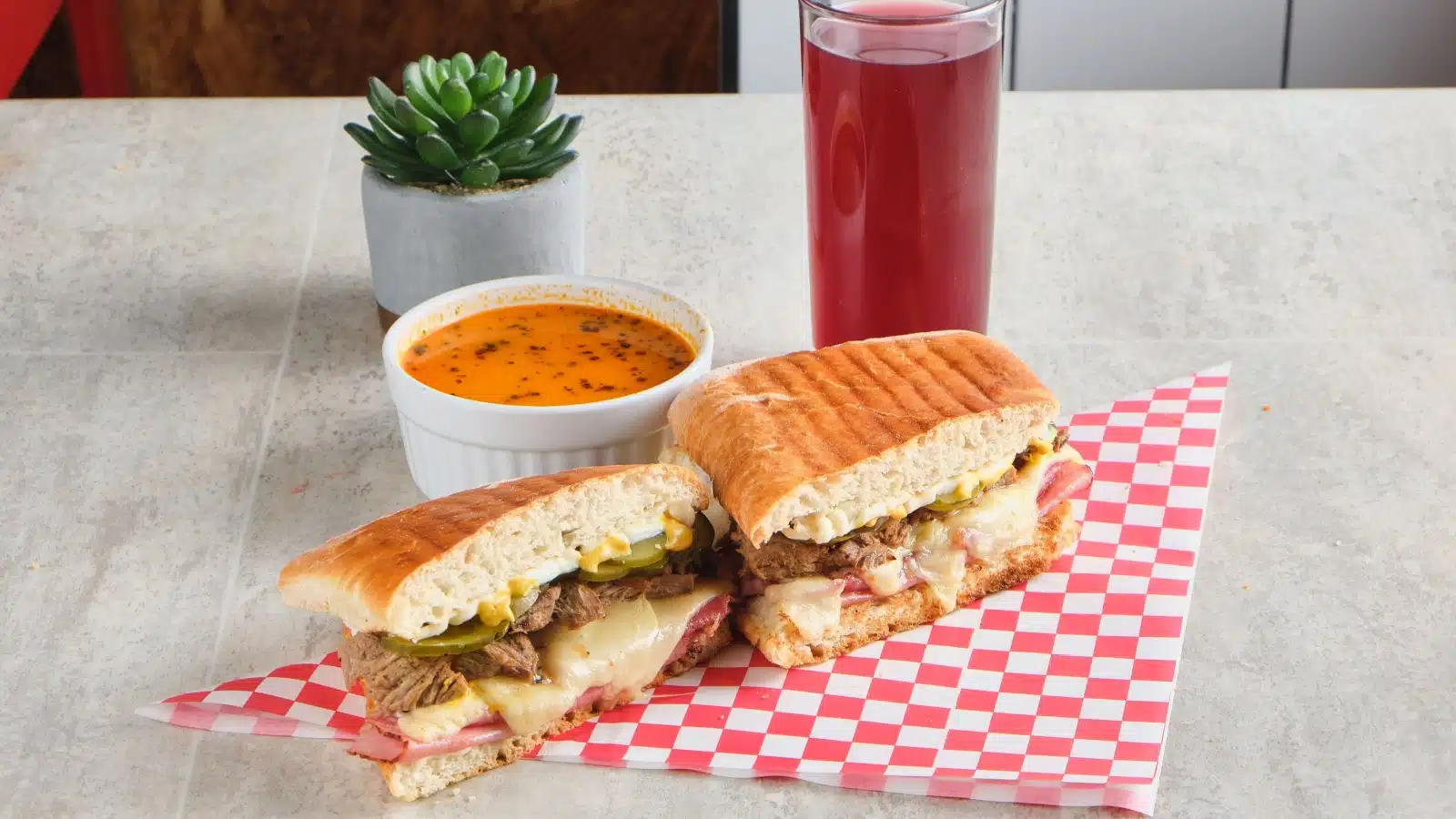 A pressed Cuban sandwich on a checkered napkin with dipping sauce and a drink.