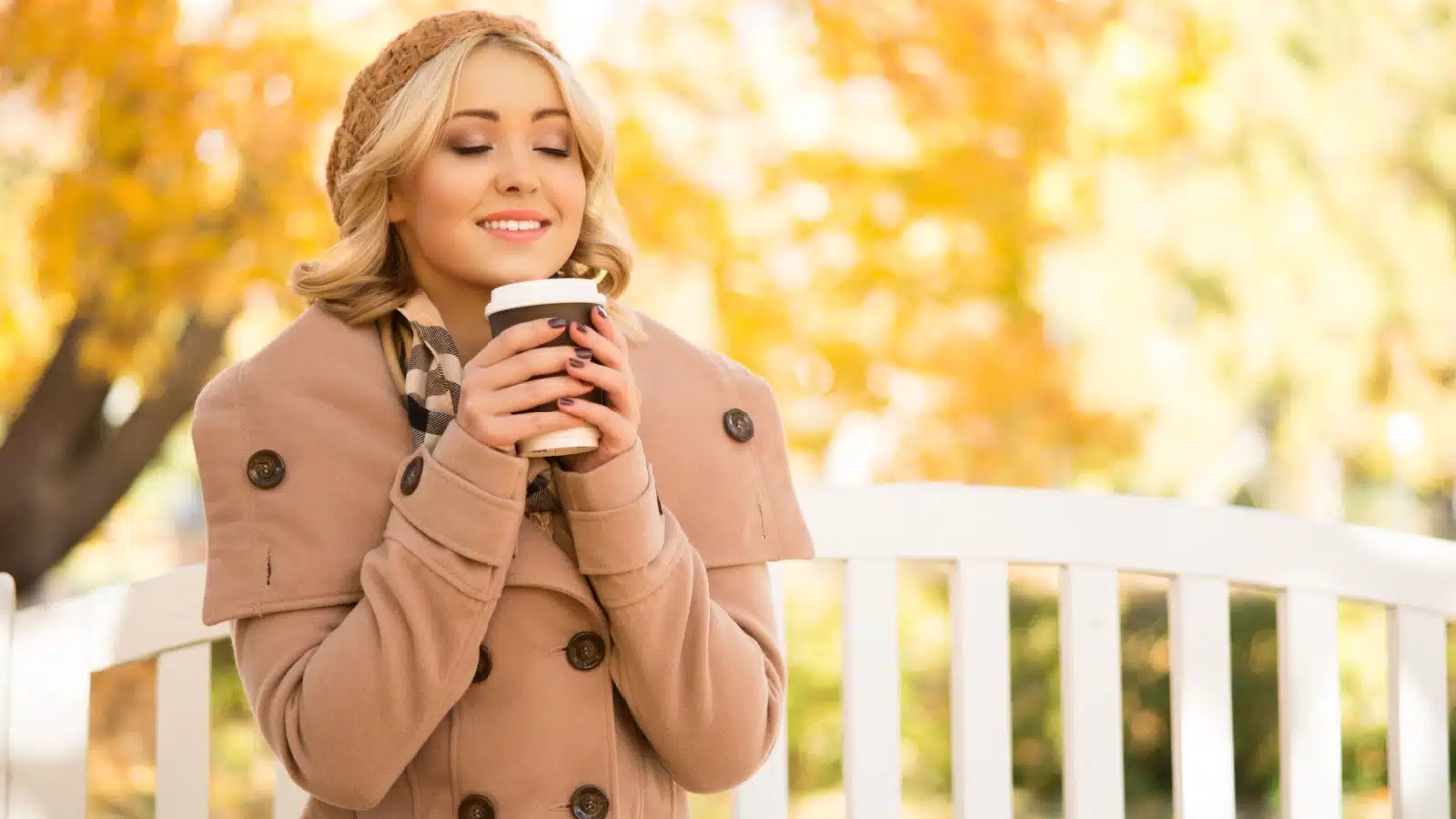 A content looking woman happily smells her hot coffee while outside on a brisk autumn day to represent fall scents.