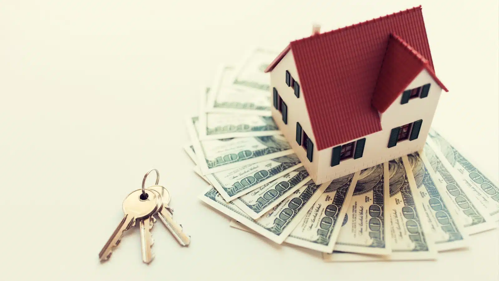 A miniature model home sits on a pile of money next to a set of keys to represent real estate closing costs.