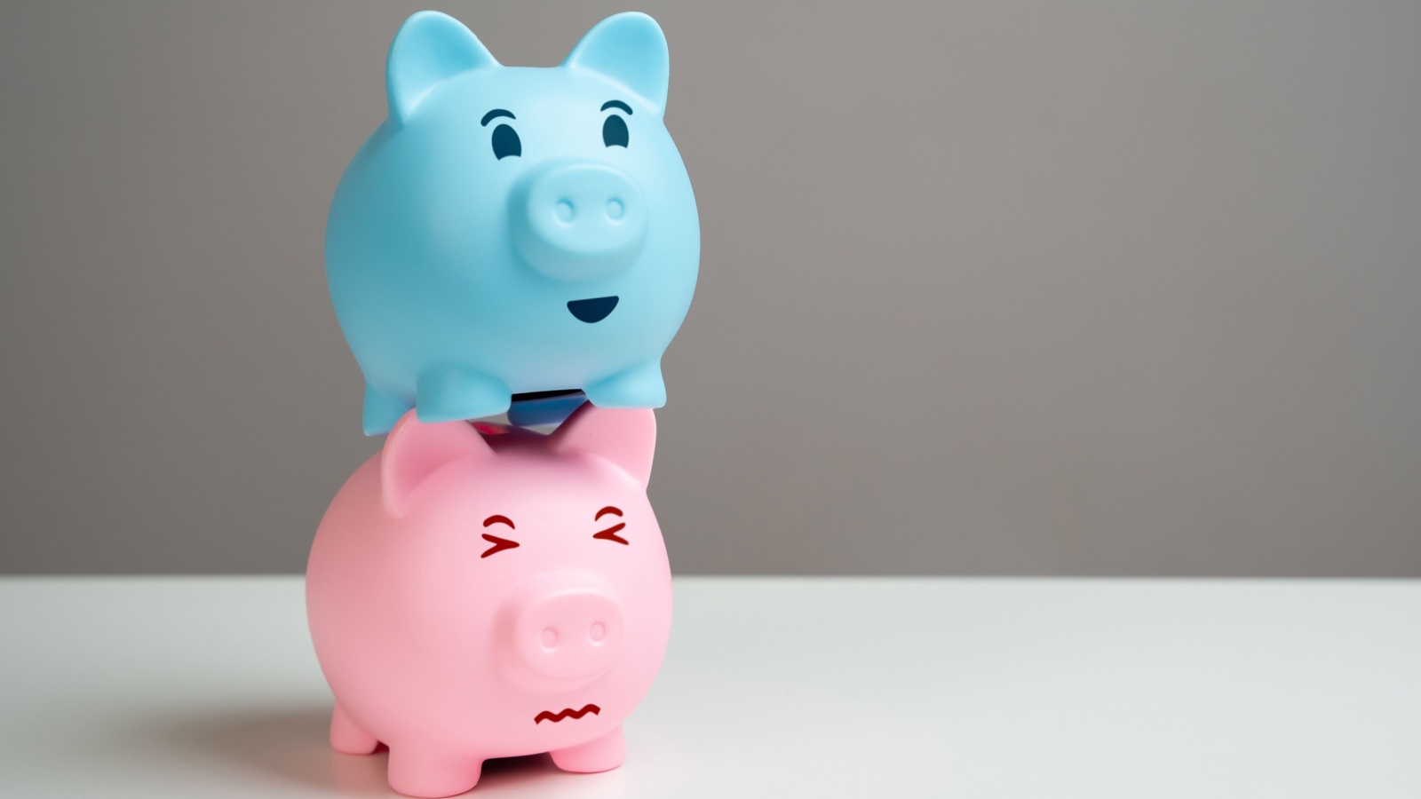 A happy blue piggy bank stands on top of a pink piggy bank, squishing it, to represent financial abuse in relationships.