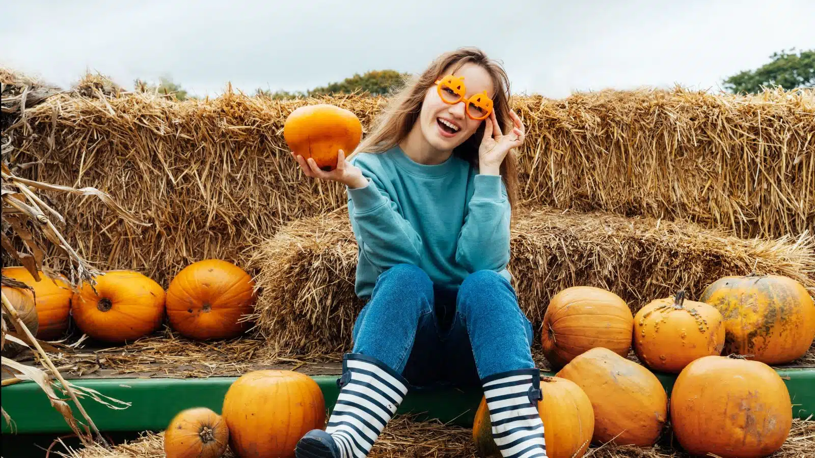A young woman smiles while holding a pumpkin and sitting on a haystack at a harvest festival.
