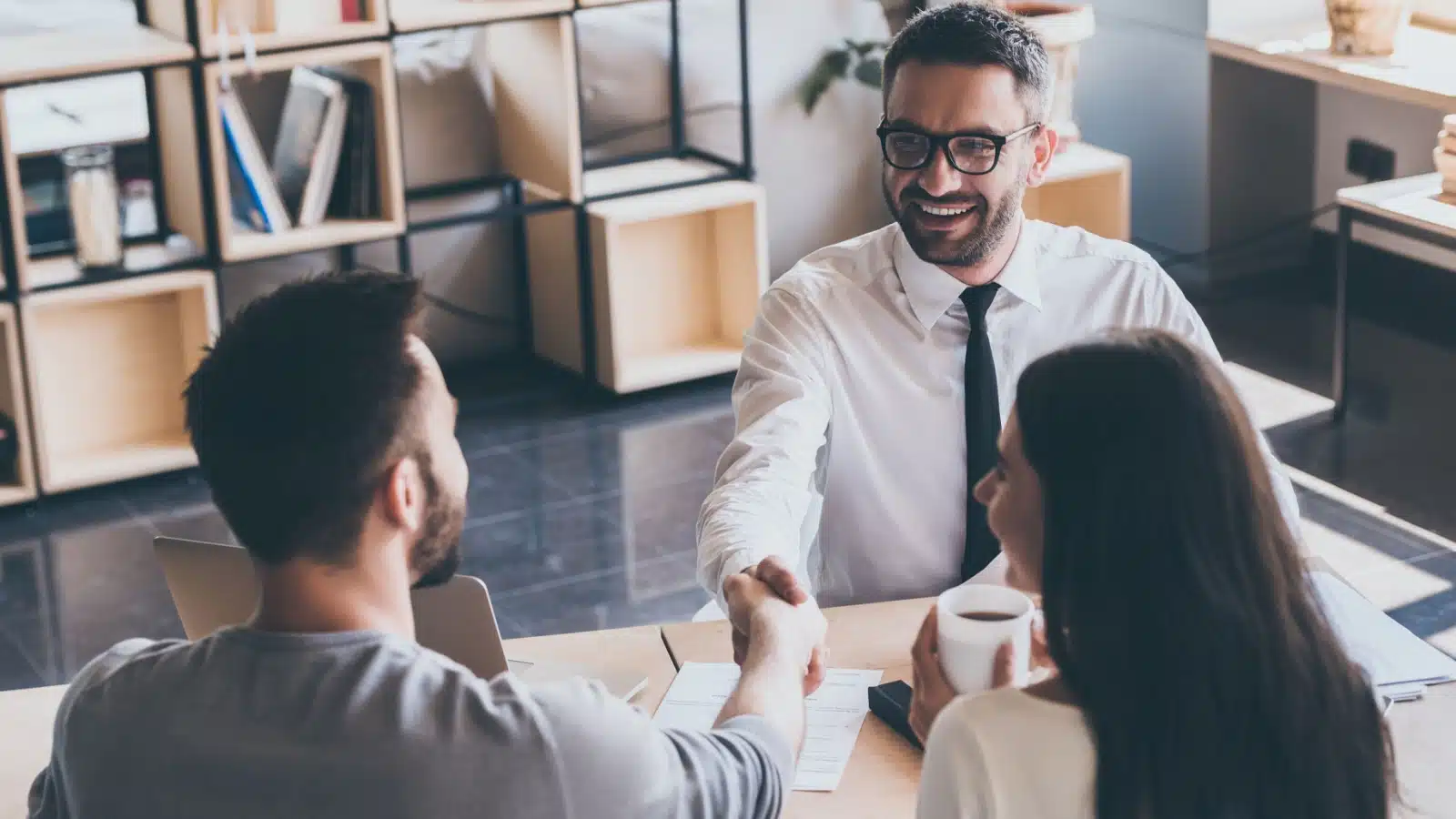 A financial advisor meets with a couple. Though he's seated across from the woman, he's focused on the man, shaking hands with him, and avoiding the woman. The image represents sexism in customer service.