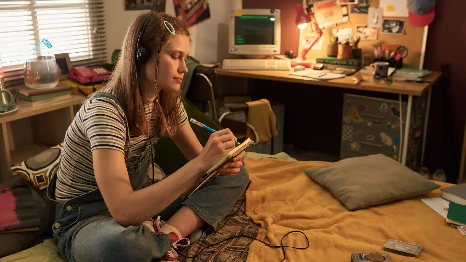 A scene from the 1990s. A girl sits on her bed journaling with an old PC on the desk behind her. She's wearing overalls. The image represents 90s nostalgia.