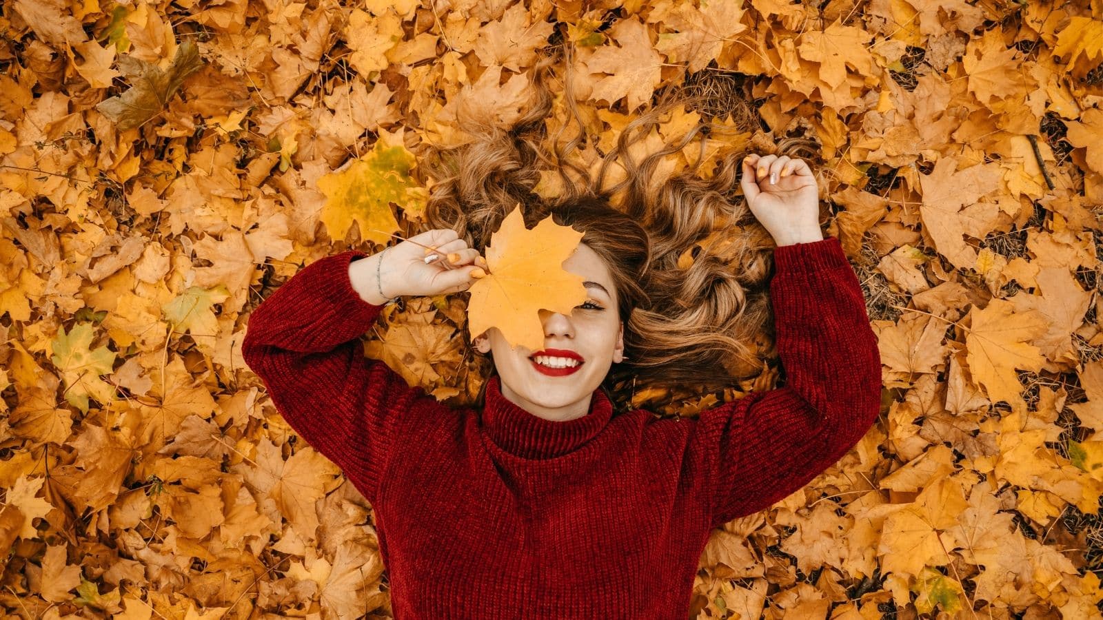 A smiling woman lies in a pile of fallen leaves, covering her eye with a giant leaf, to represent cheap fall activities.