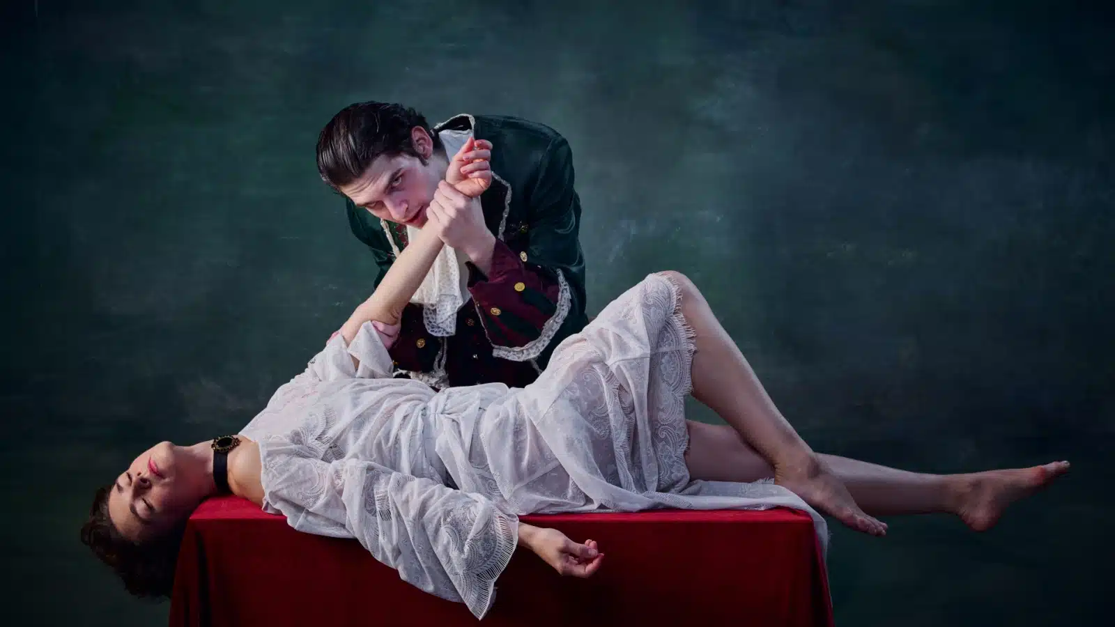 A male vampire stands over his female victim, holding her arm up to feed. She's laying, unconscious, on a small table. She's dressed in white to represent purity, while he's clothed in mostly black.