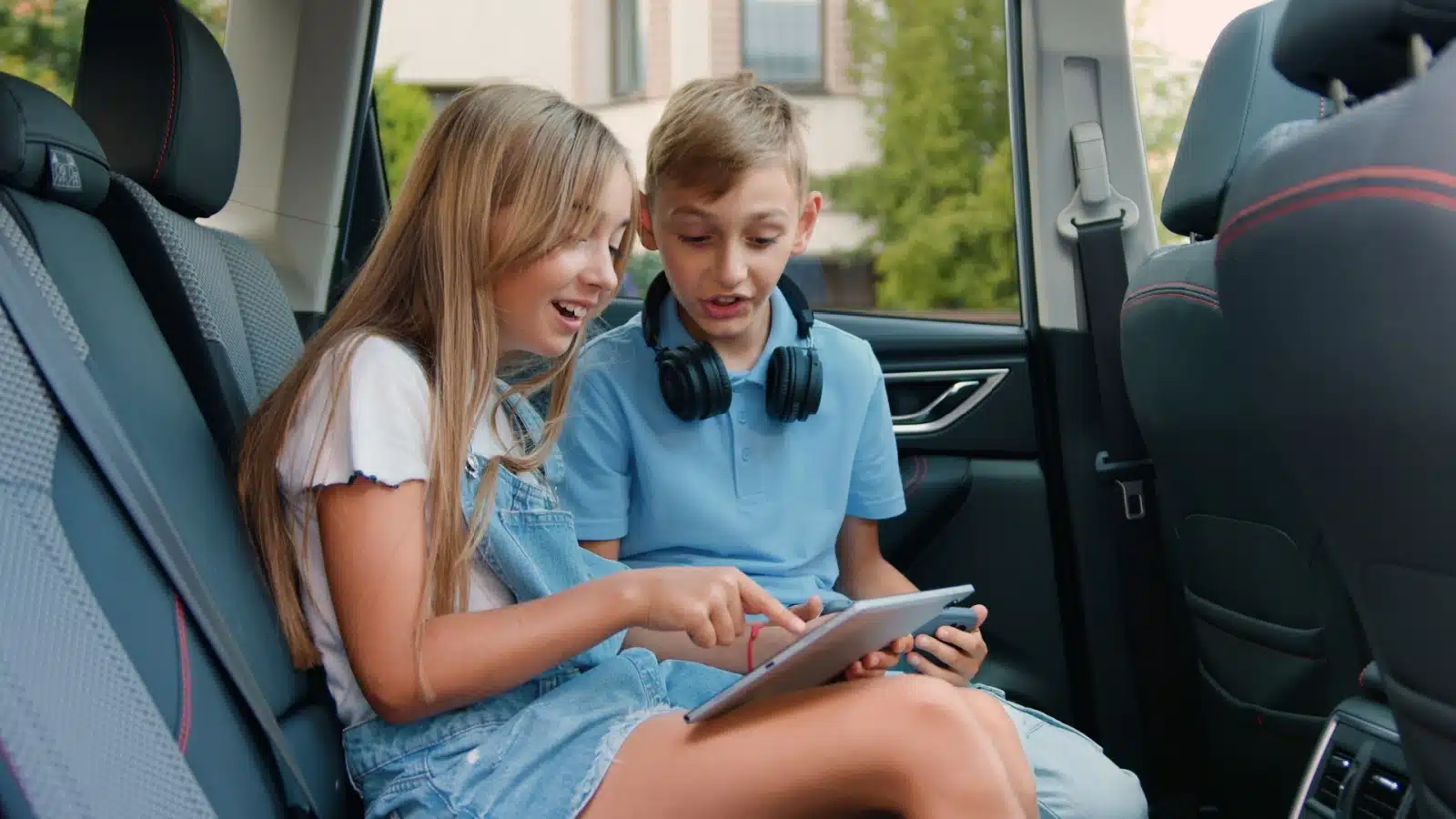 Kids playing road trip games on a tablet in the back seat of a car.