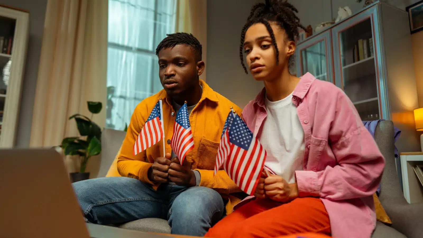 A couple sits on the couch holding mini American flags while sadly watching the news to represent the biggest problems in America.
