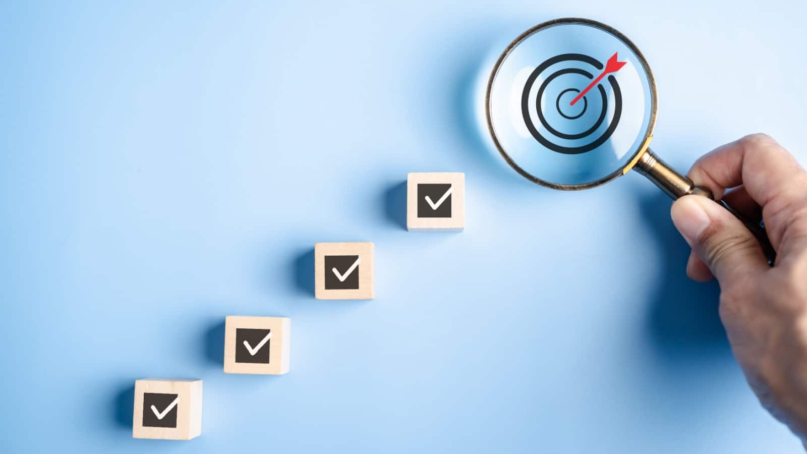 A hand holds a magnifying glass over a target. Little wooden blocks with checkmarks on them lead up to the target, to represent steps to achieve in a process goal.