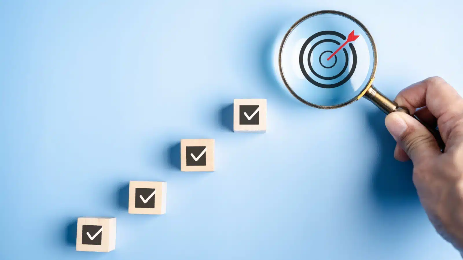A hand holds a magnifying glass over a target. Little wooden blocks with checkmarks on them lead up to the target, to represent steps to achieve in a process goal.