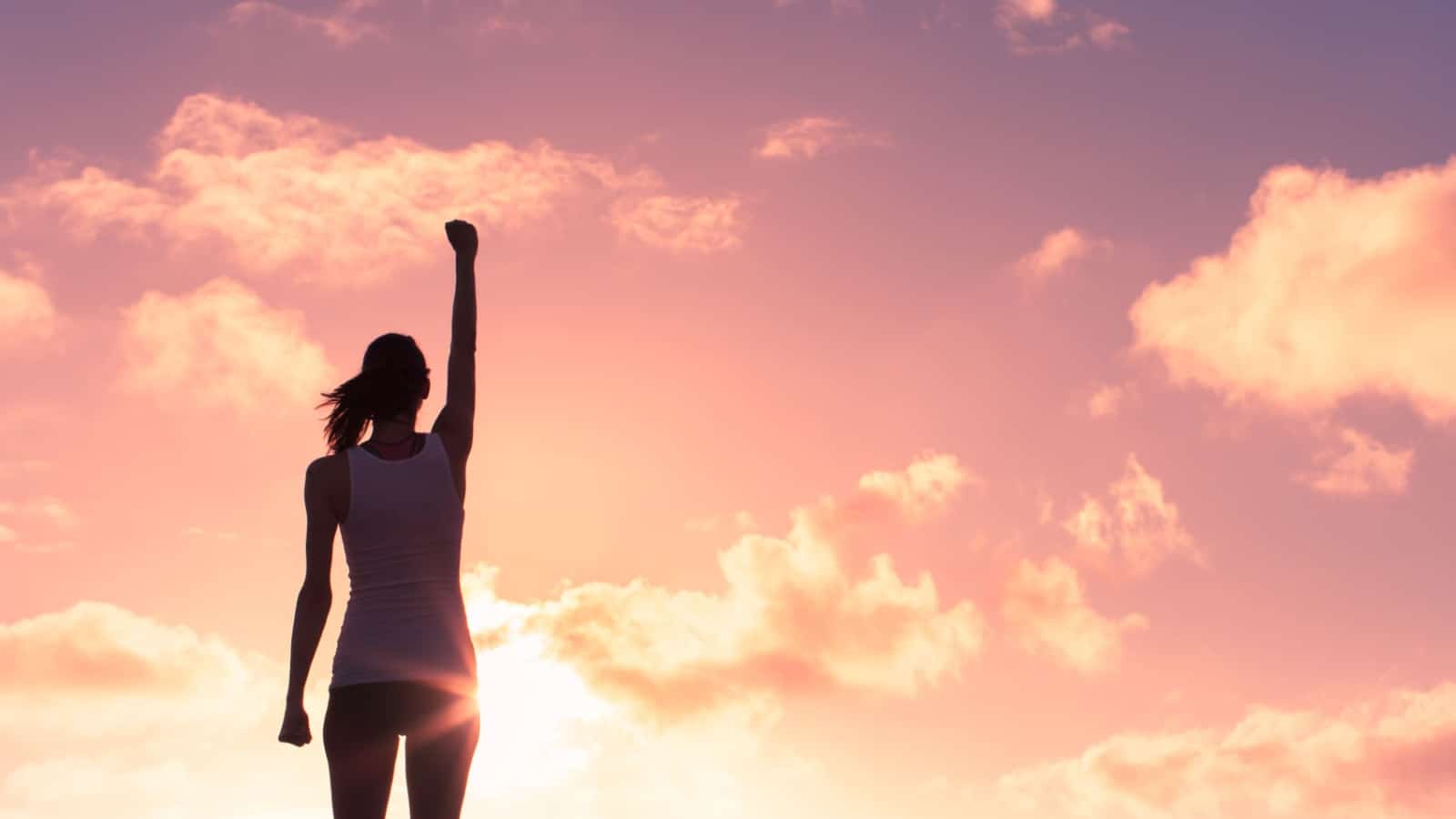 Silhouette of woman raising her fist in the air at sunset to represent self-improvement.
