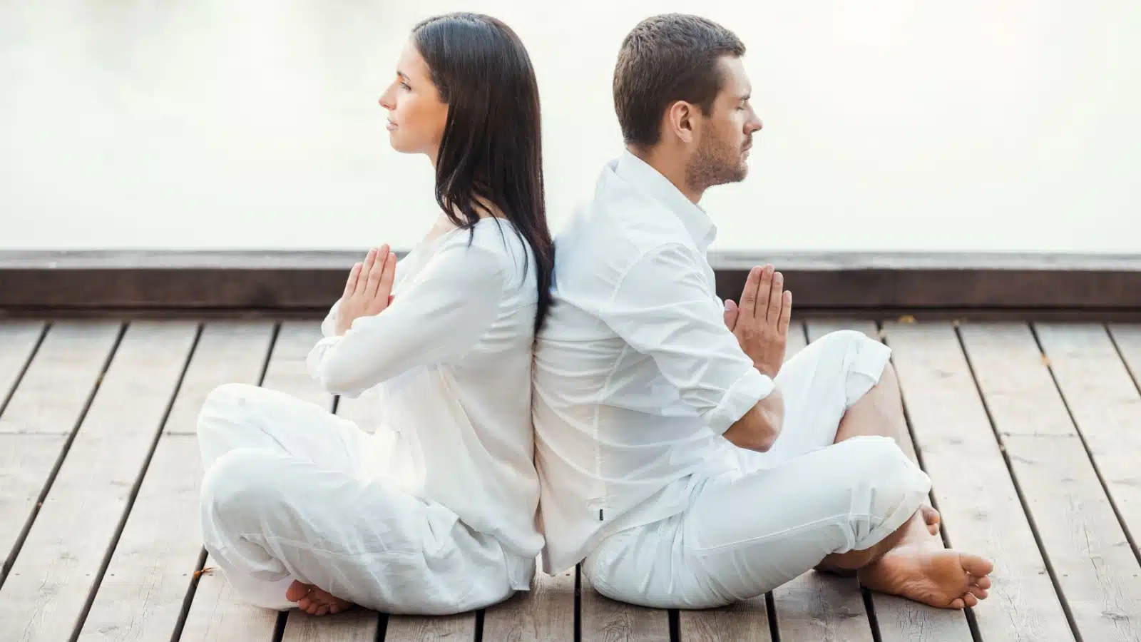 A man and women, both dressed in all white, sit back to back in a balanced meditation.