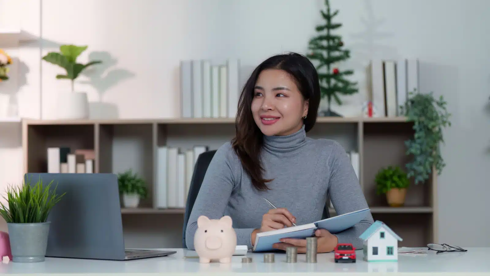 A woman considers her financial goals. There's a piggy bank, stack of change, model car, and model house in front of her to represent different goals.